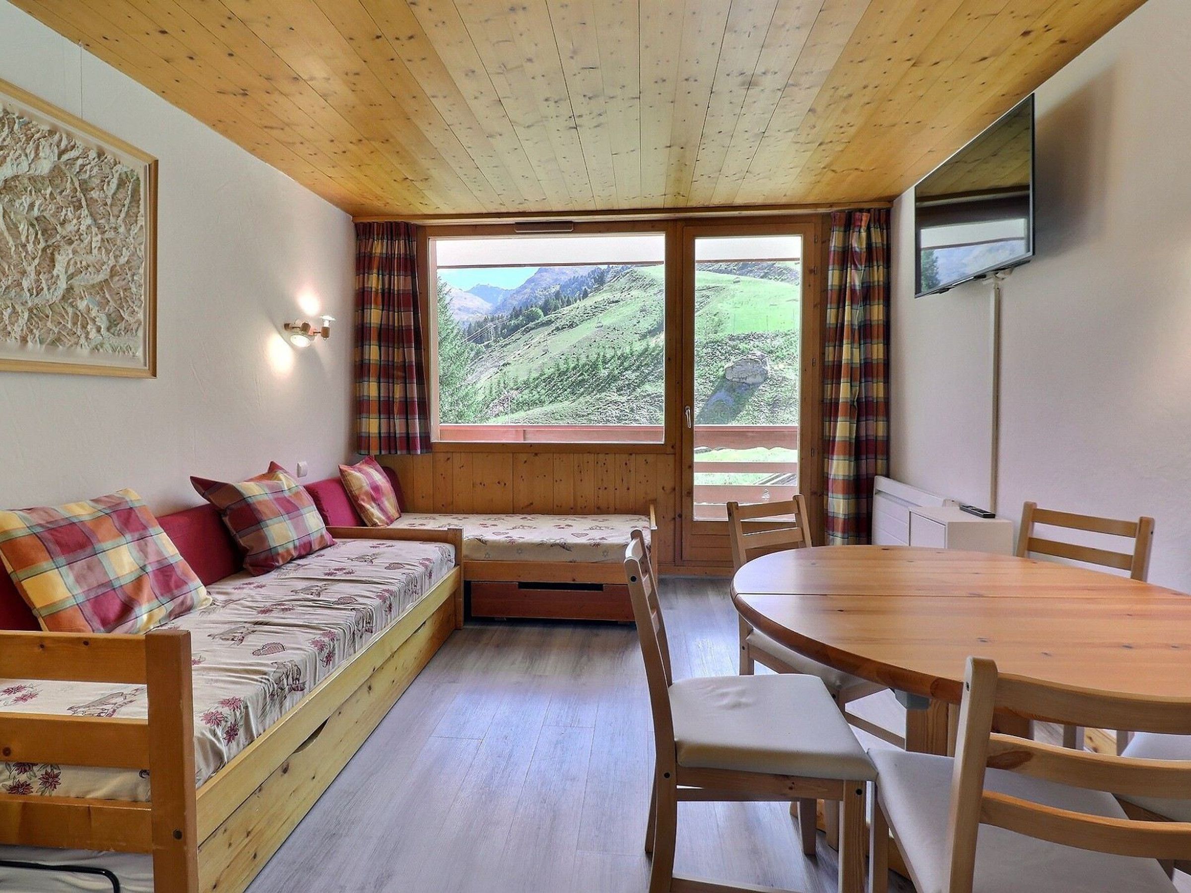 A cozy cabin with a wooden interior, a sofa with plaid cushions, a dining table, and a view of a green valley.