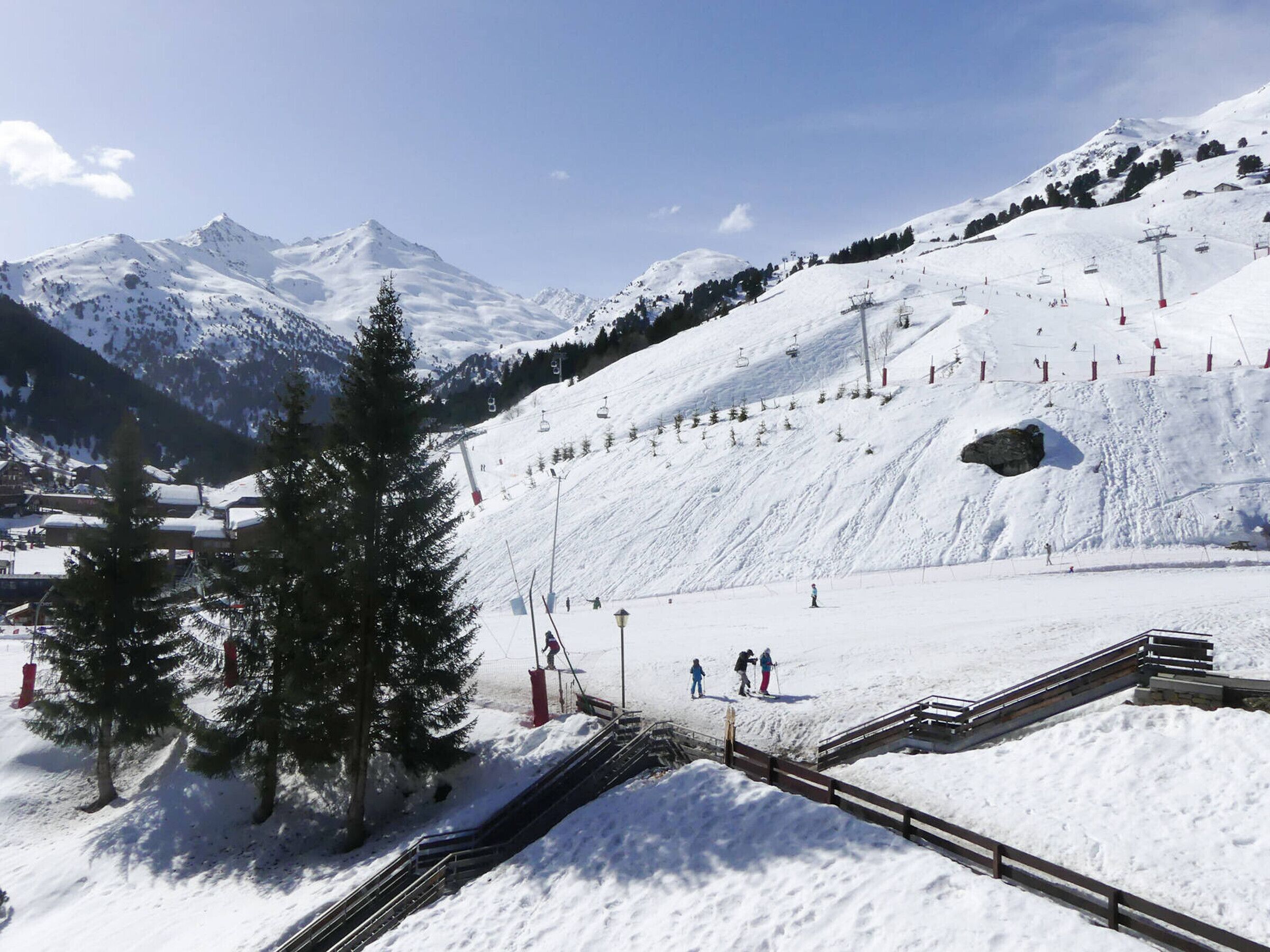 A snow-covered ski slope with ski lifts, a wooden viewing platform, and a mountain backdrop.
