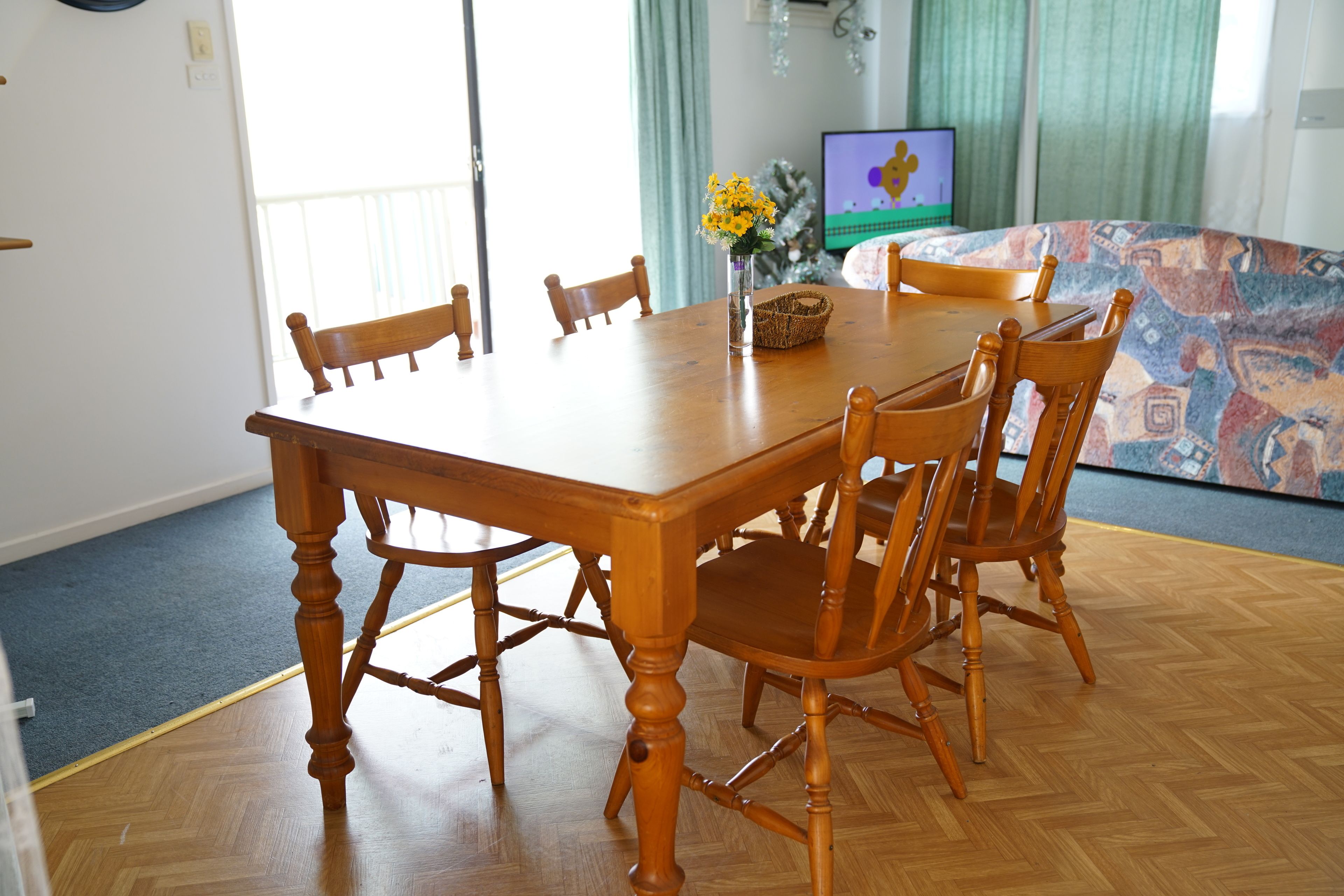 Een eetgedeelte met een houten tafel en stoelen, een vaas met gele bloemen, en een televisie op de achtergrond.