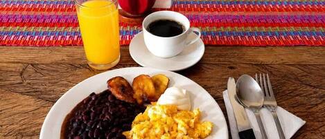 A breakfast plate with beans, scrambled eggs, and plantains, a glass of orange juice, and a cup of black coffee on a wooden table with a colorful woven placemat.