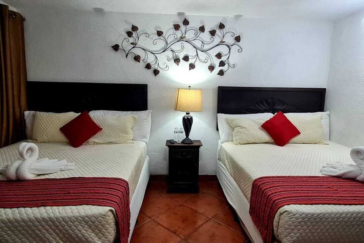 A hotel room with two beds, each with a red pillow and a white towel. There is a wall-mounted light fixture above the beds and a small table between them.