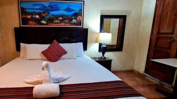 Habitación de hotel con cama, un cuadro en la pared, mesita de noche con lámpara y puerta de madera.