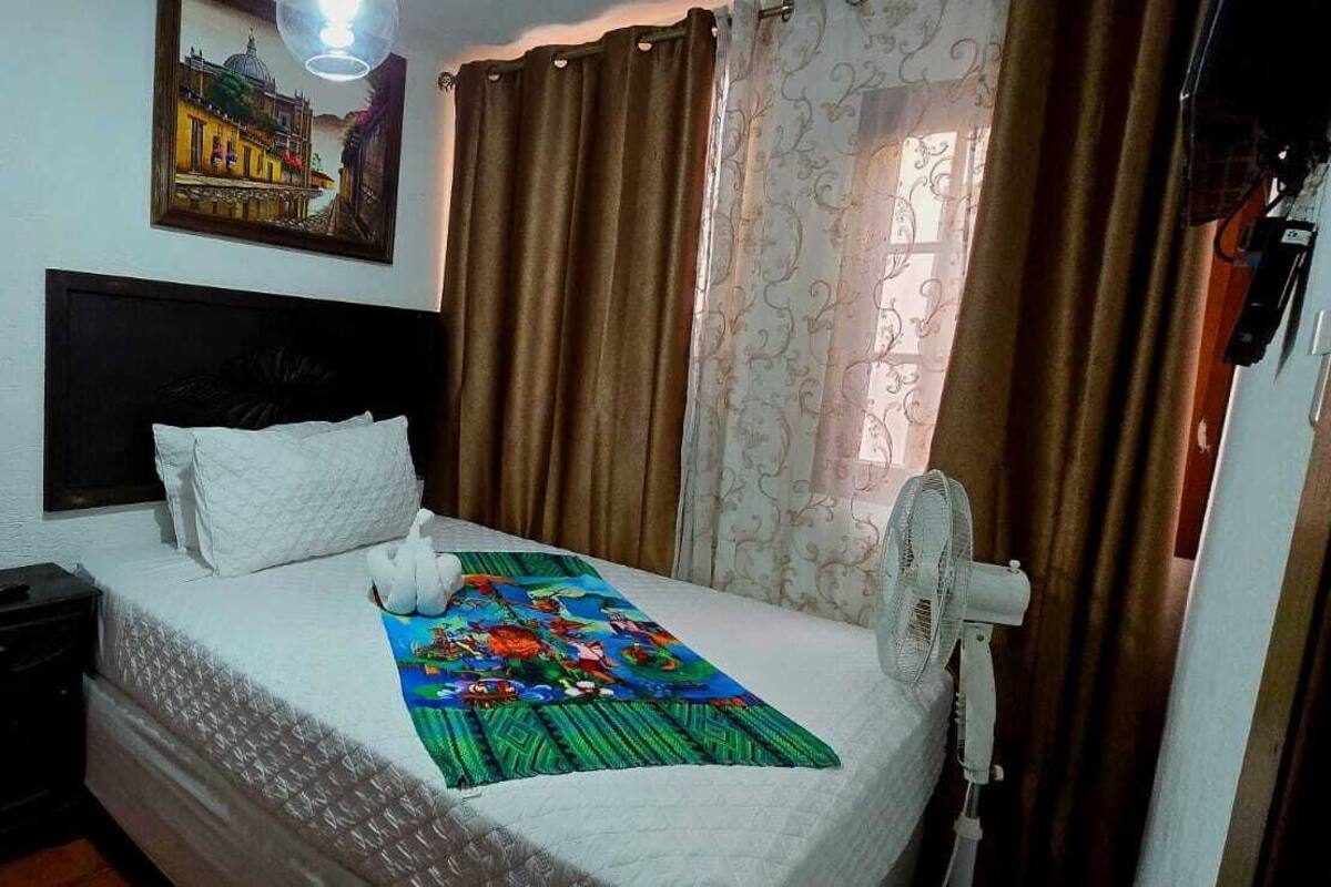A bedroom with a bed, a nightstand, a bedside table, a wall-mounted fan, and a window with curtains.