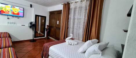 A hotel room with a bed, a television, a fan, and a door.