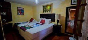 Una habitación de hotel con una cama, una mesita de noche, una lámpara, un ventilador y dos cuadros en la pared.