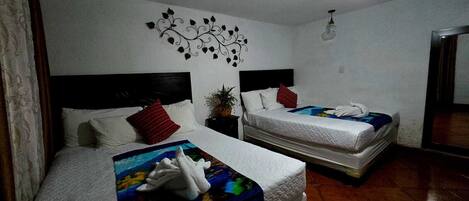Habitación de hotel con dos camas, cada una con un colchón blanco y una manta colorida. Encima de las camas hay un adorno de pared y una pequeña mesa con una planta.
