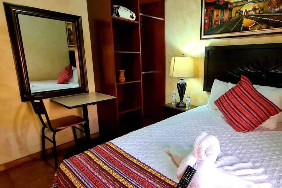 A hotel room with a bed, a desk, a mirror, a wardrobe, and a painting on the wall.