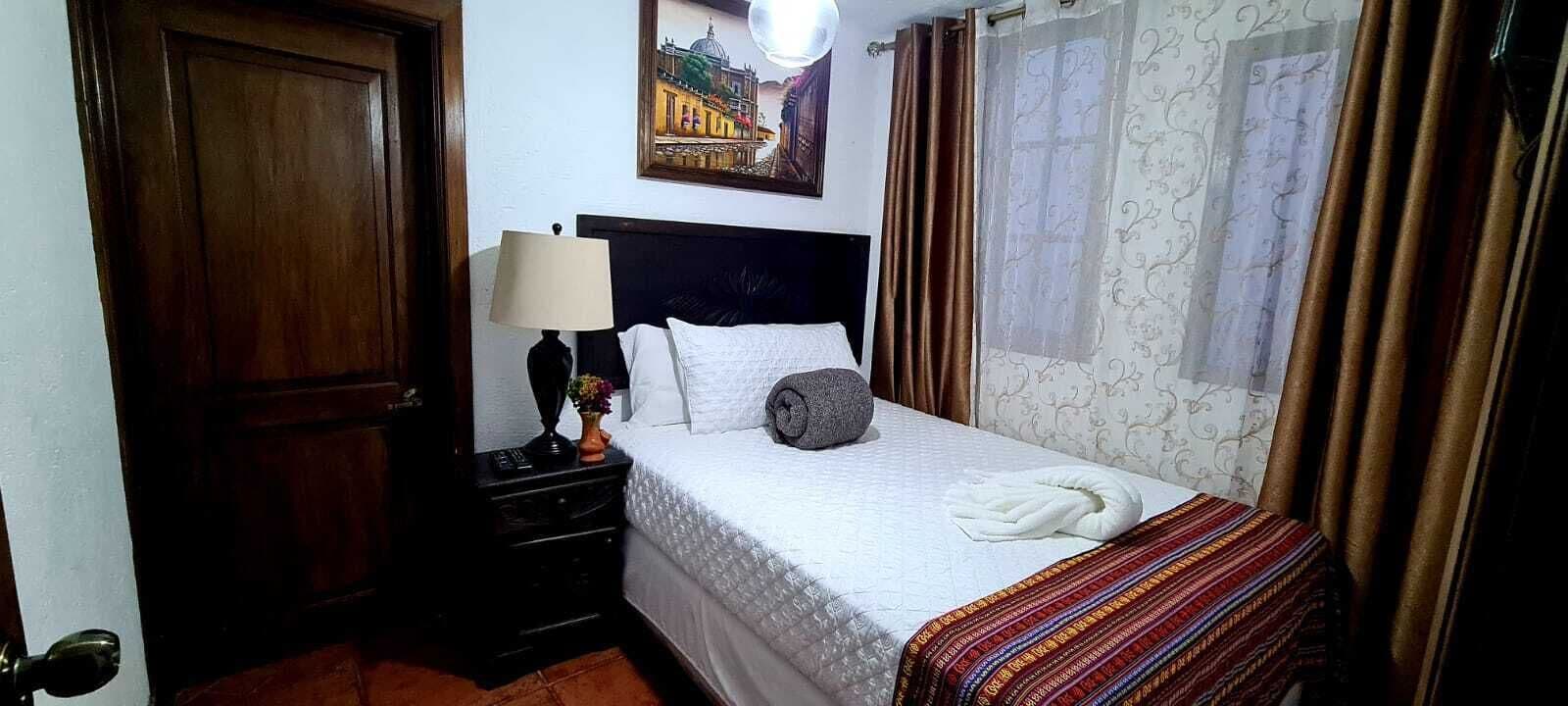 A hotel room with a bed, a nightstand, a lamp, a window with curtains, and a wall-mounted painting.