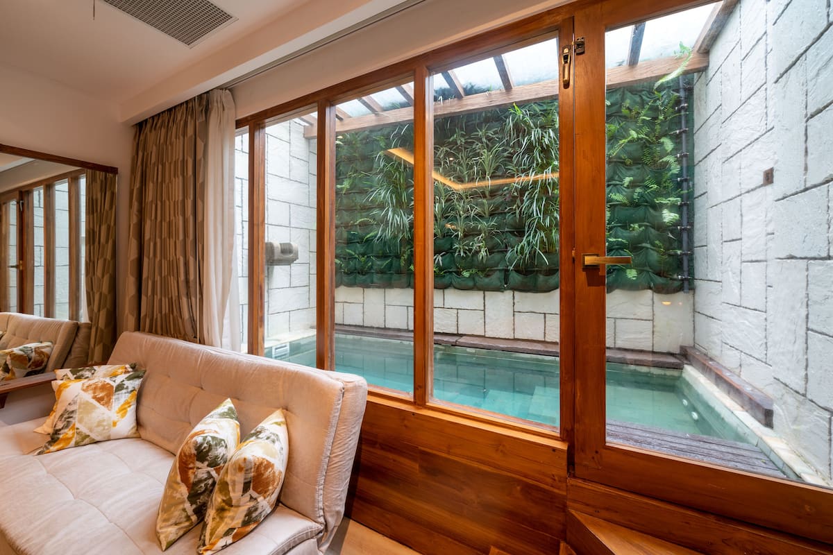 Ein Zimmer mit einer hölzernen Bank, einem Sofa mit gemusterten Kissen und großen Fenstern mit Blick auf einen Poolbereich mit viel Grün.