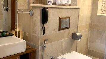 A modern bathroom with a white sink, toilet, and shower. There are towels on a shelf and a phone mounted on the wall.