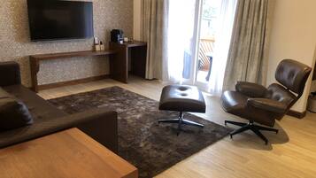 A modern living room with a flat-screen TV, a wooden coffee table, a brown leather sofa, a leather armchair, and a dark rug.