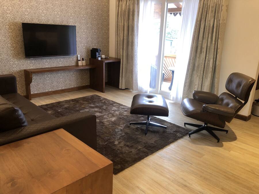 A modern living room with a flat-screen TV, a wooden coffee table, a brown leather sofa, a leather armchair, and a dark rug.