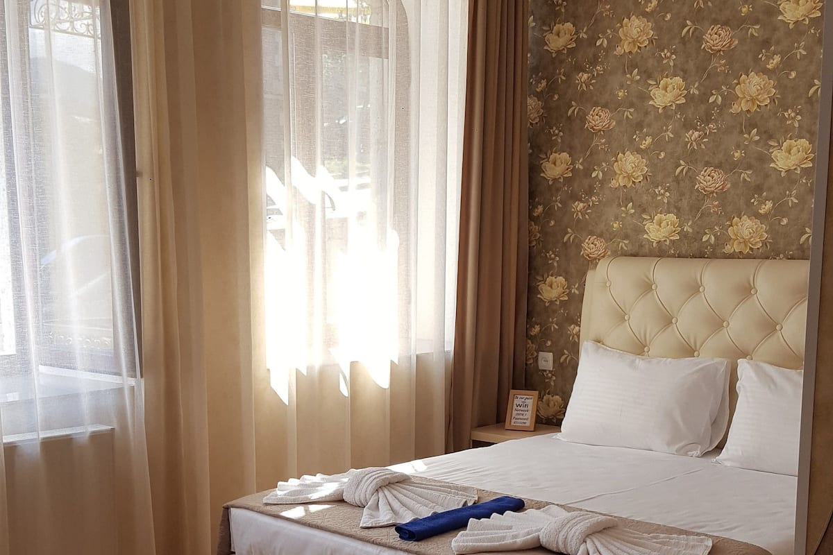 Ein Hotelzimmer mit einem großen Bett, Holzboden und Blick ins Freie durch transparente Vorhänge.