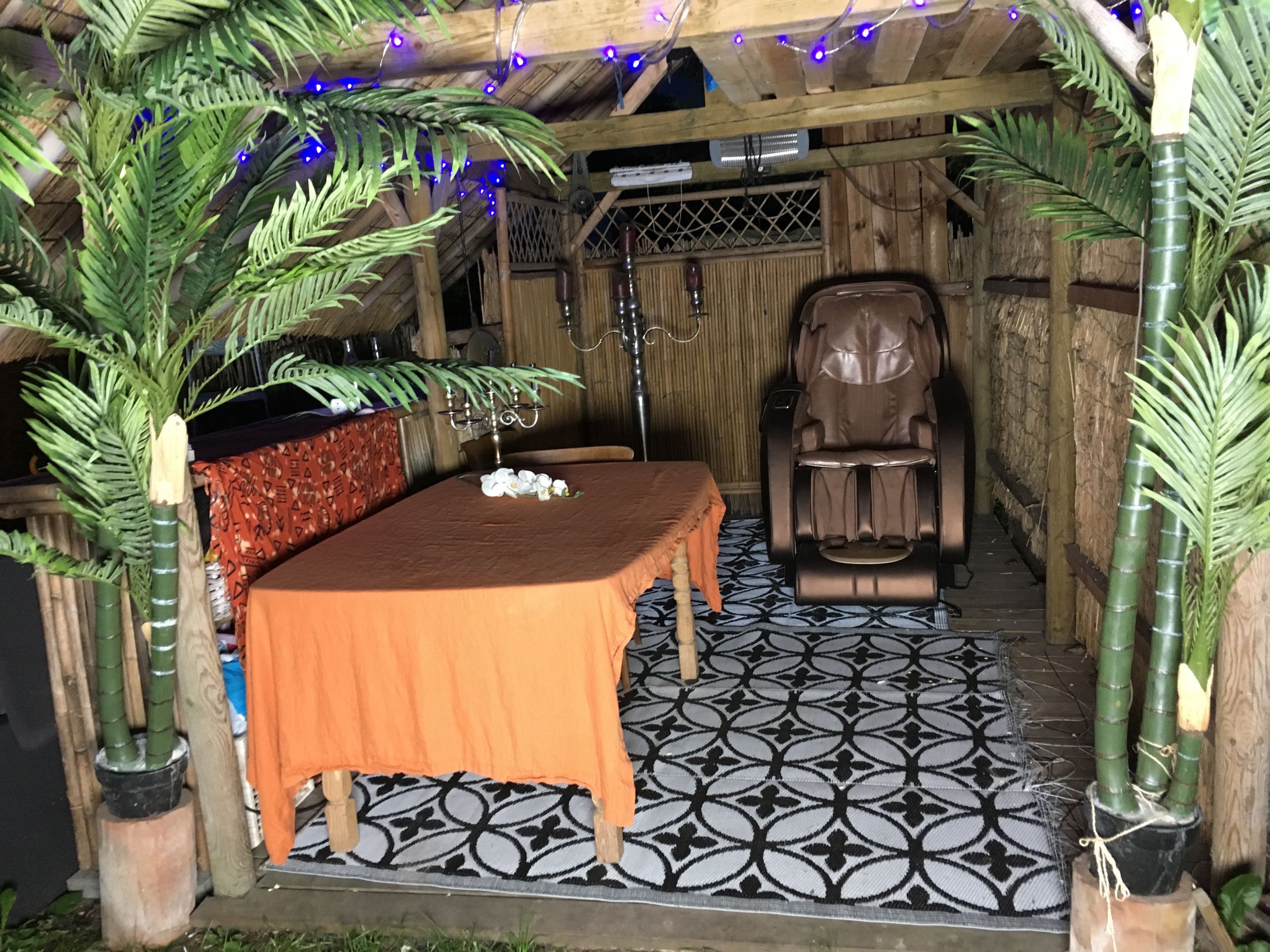 A massage chair in a room with a patterned floor, a table with a blanket, and decorative plants.
