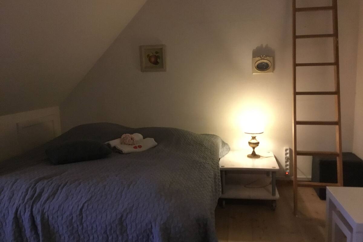 Makuuhuoneessa on sänky, yöpöytä, lamppu, tikkaat ja seinällä oleva kehystetty kuva.