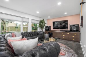 A modern living room with a sectional sofa, a flat-screen TV, a sound system, and a large window with blinds. - Experience Massapequa charm near Roosevelt Field Mall, 15 Mins to Jones Beach (Massapequa)