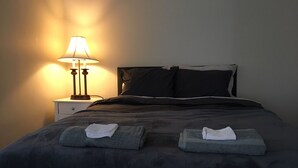 Una cama bien hecha con un edredón oscuro y almohadas, una lámpara de noche y dos toallas dobladas sobre la cama.