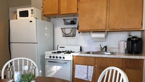 Cocina compacta con armarios de madera, un refrigerador blanco, estufa y microondas. Hay un lavamanos con grifo, una cafetera y una pequeña planta en maceta sobre la mesa.