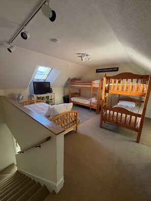 Ein Schlafsaalzimmer mit Etagenbetten, einem Schreibtisch mit Computer, einem kleinen Fenster und einem Deckenventilator.