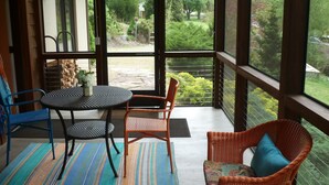 Una habitación con puerta de vidrio que da a un jardín, una mesa con sillas y un sillón de mimbre con cojín.