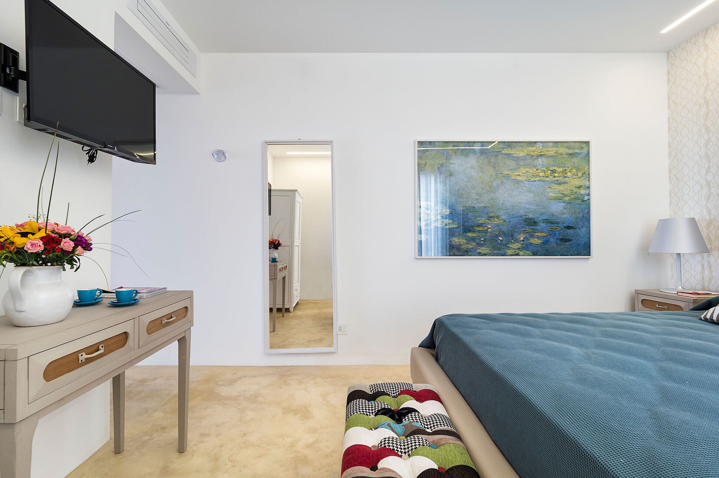 A bedroom with a bed, a desk with a vase of flowers, a painting, and a mirror.