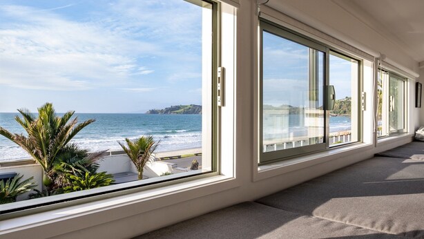 A room with large windows overlooking a beach, a sofa, and a small plant. - Absolute beachfront position on Waiheke Island. (Onetangi)