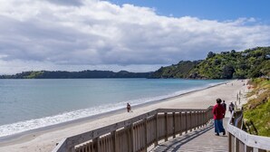 A wooden boardwalk leading to a sandy beach with a view of the sea and distant hills. - Absolute beachfront position on Waiheke Island. (Onetangi)
