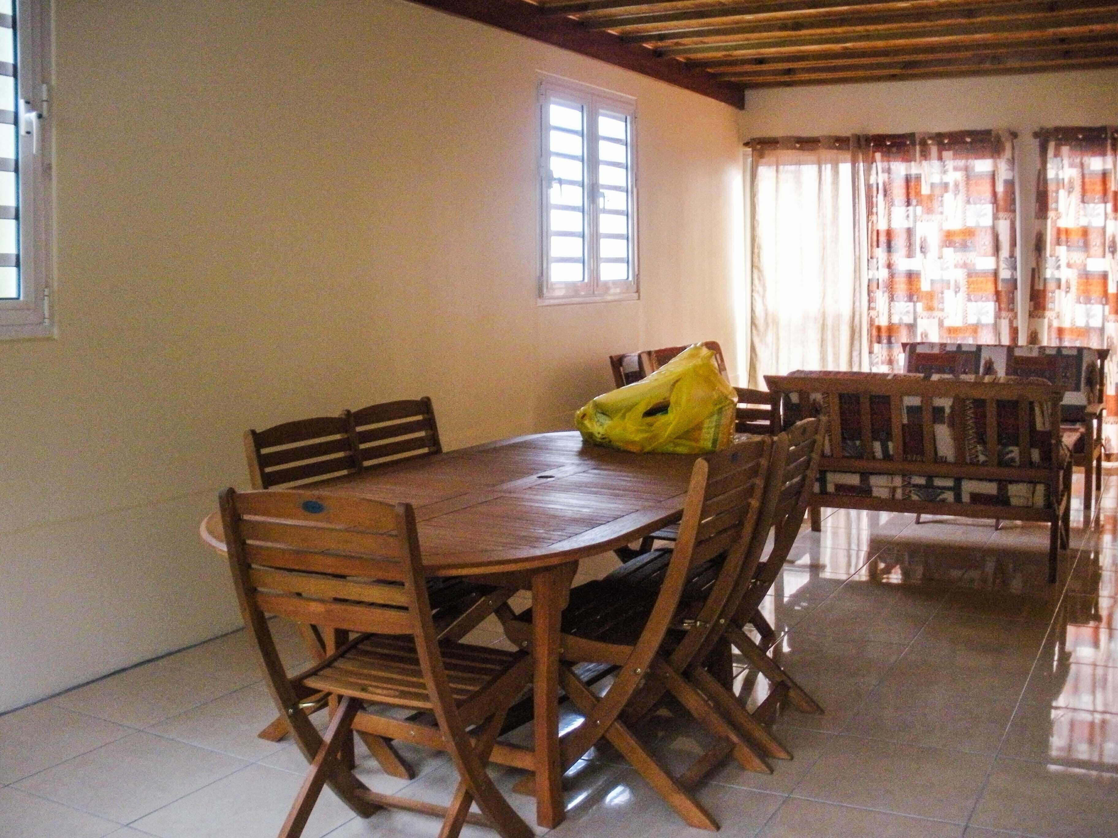 A dining area with a wooden table and chairs, a yellow bag on the table, and a window with blinds.