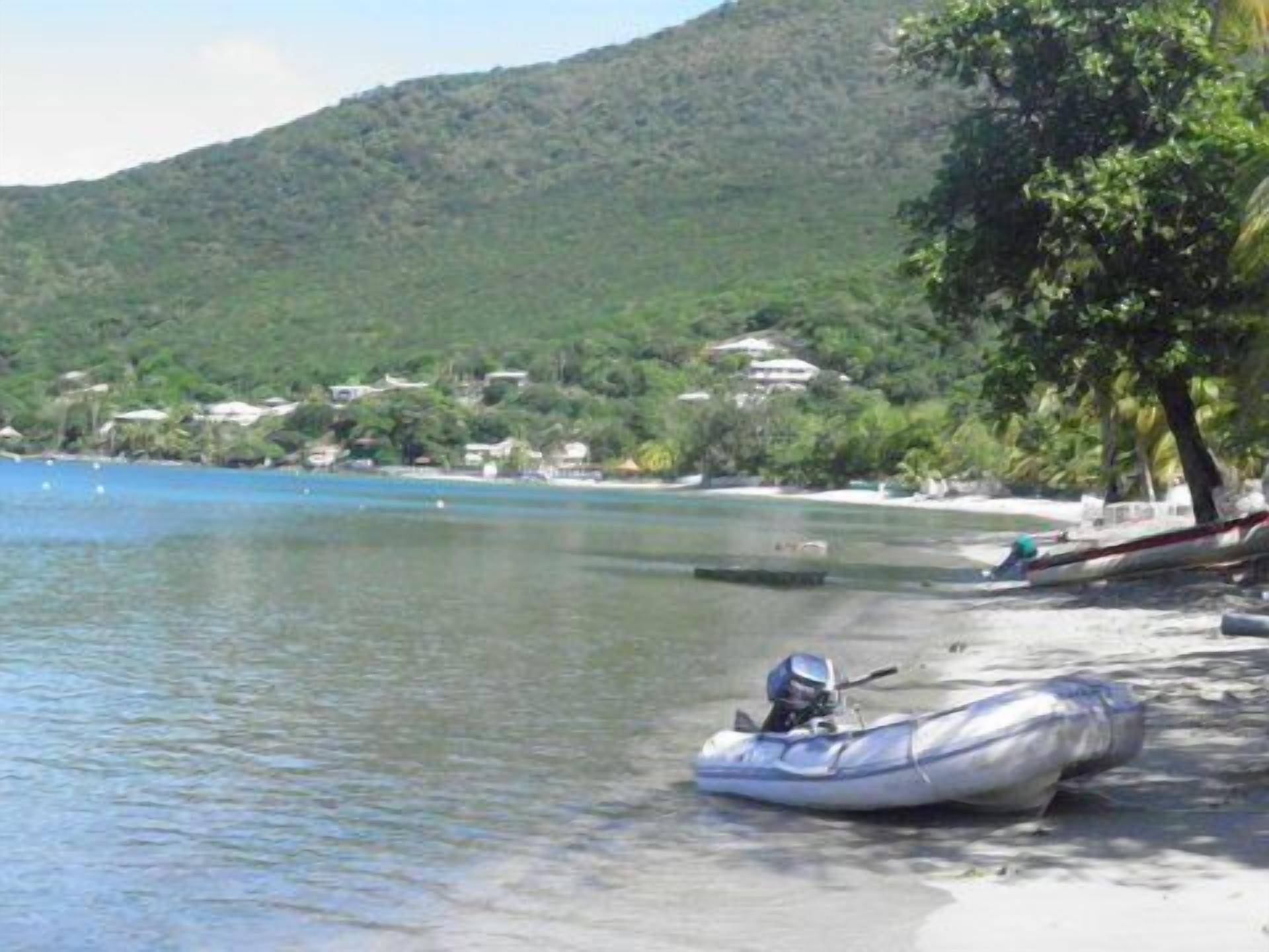 A beach with a clear blue sea, a small boat on the shore, and a hill with buildings in the background.