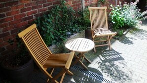 Zwei Holzstühle und ein kleiner runder Tisch auf einer Terrasse mit Backsteinwand und Pflanzkübeln.