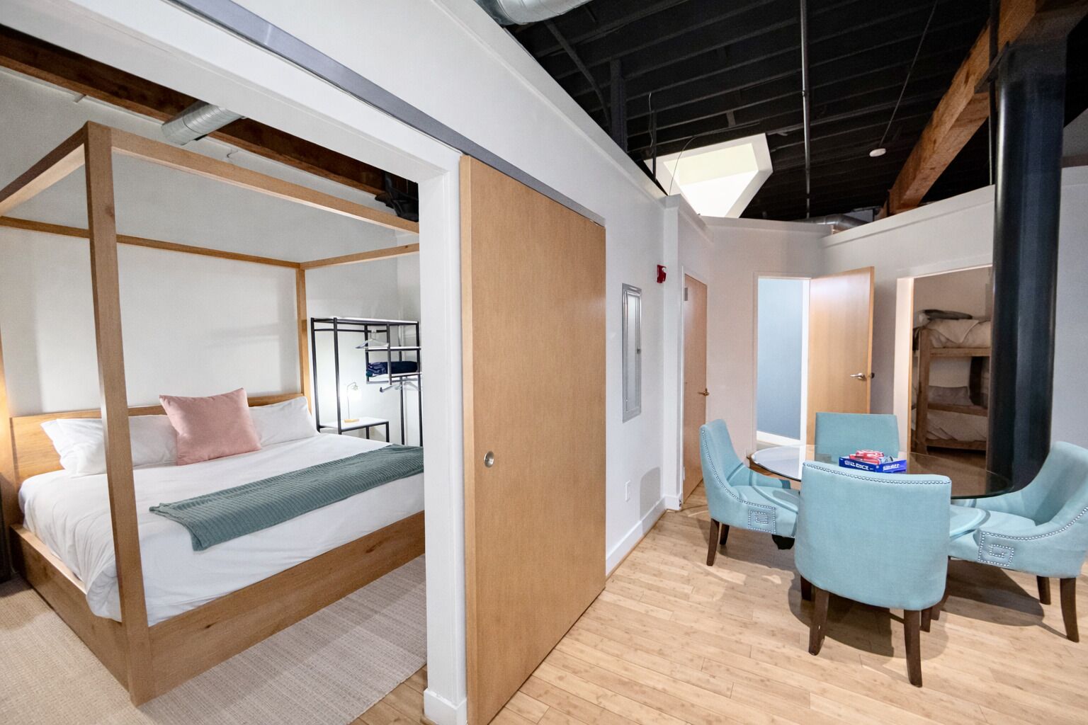 A modern hotel room with a four-poster bed, a wooden floor, a dining area with two light blue chairs, and a bunk bed in the background.