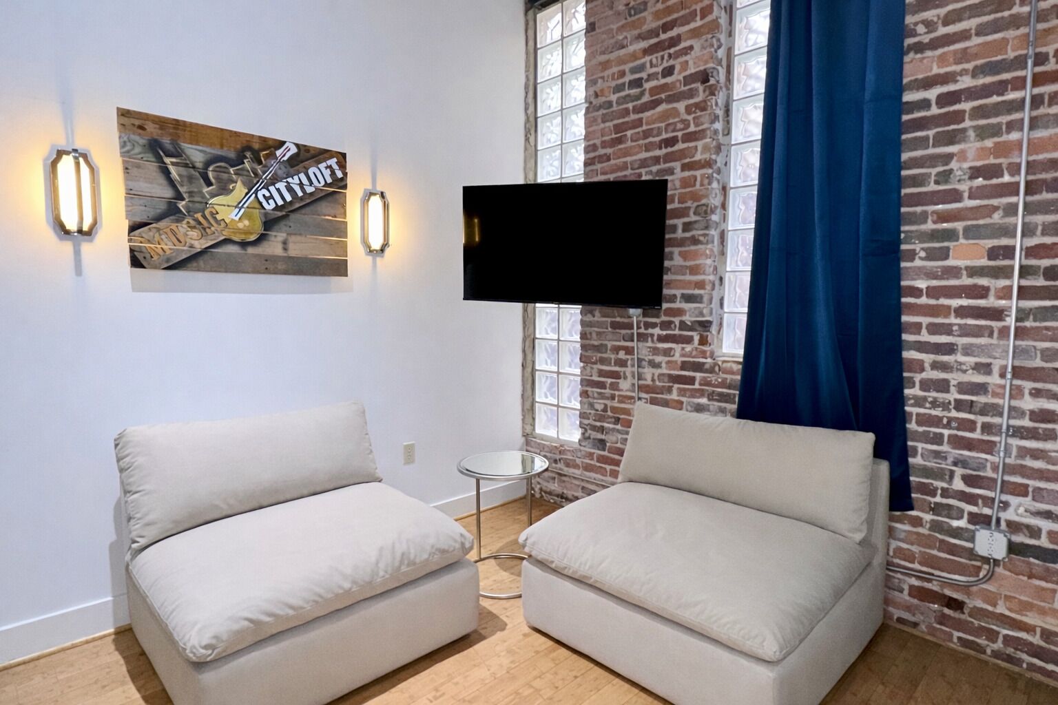 Una habitación con dos sofás beige, un televisor de pantalla plana montado en la pared de ladrillo y una pequeña mesa redonda.