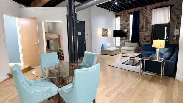 A spacious living area with a glass dining table surrounded by four chairs, a sofa set, a coffee table, and a TV mounted on a brick wall. - Downtown Music City Loft living! ~Steps to the Ryman and Broadway (Nashville)