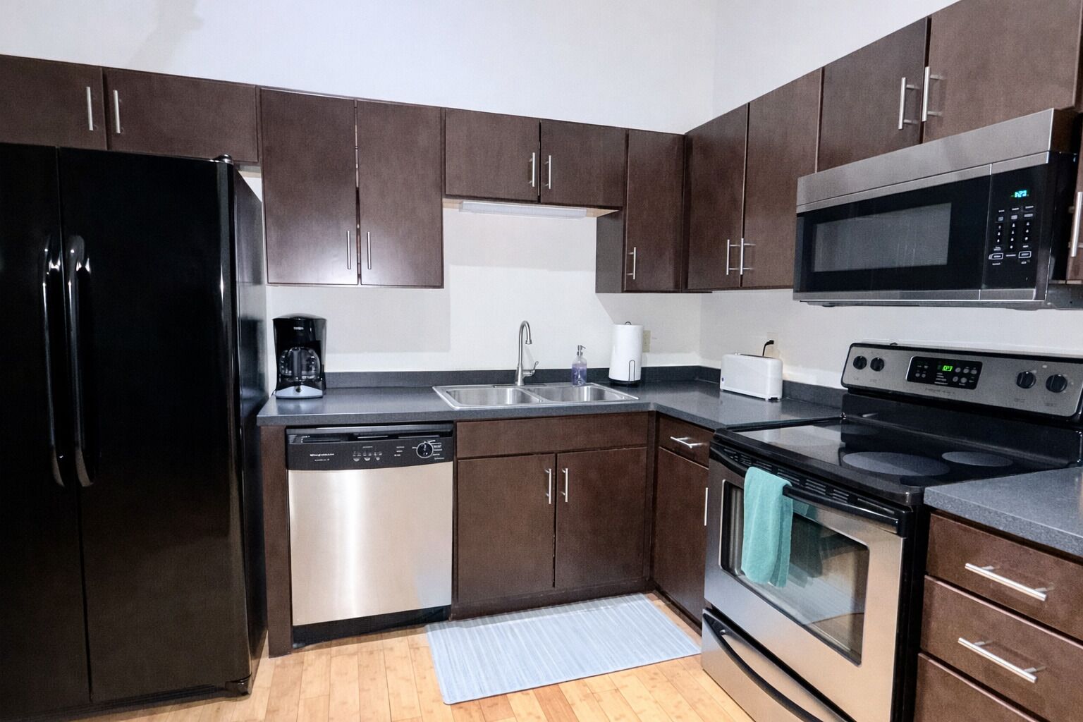 Eine moderne Küche mit dunkelbraunen Schränken, Edelstahlgeräten, darunter ein Kühlschrank, eine Spülmaschine, eine Mikrowelle und ein Herd, sowie einer grauen Arbeitsplatte.