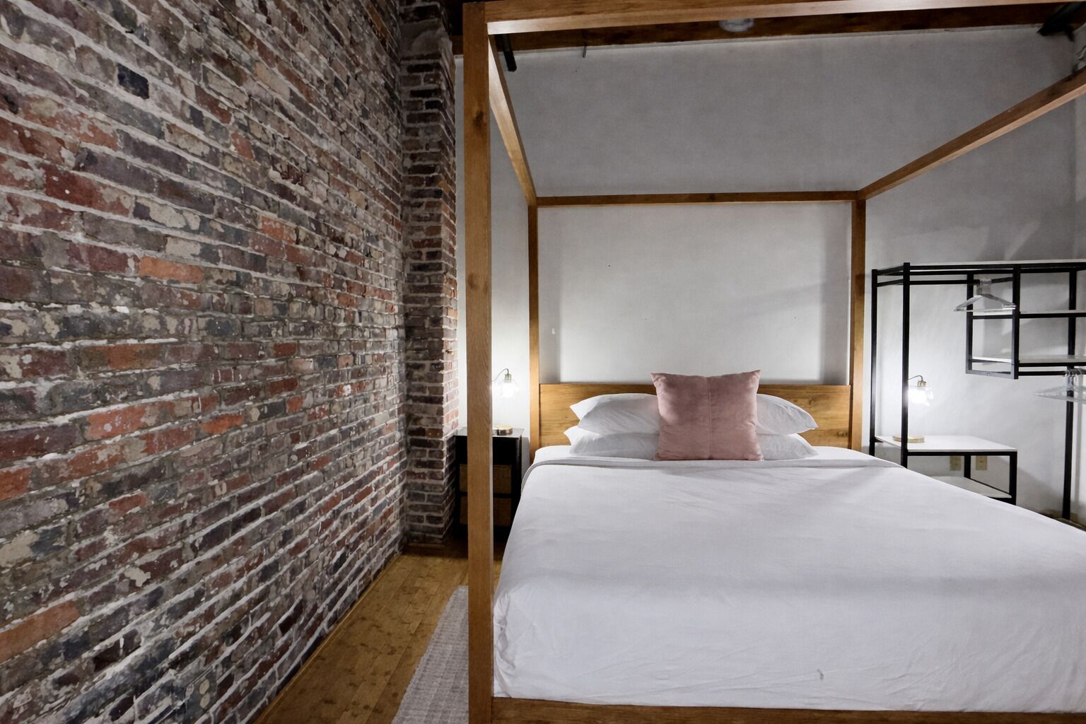 A bedroom with a brick wall, a wooden headboard, a white bed with a pink pillow, and a nightstand with a lamp.