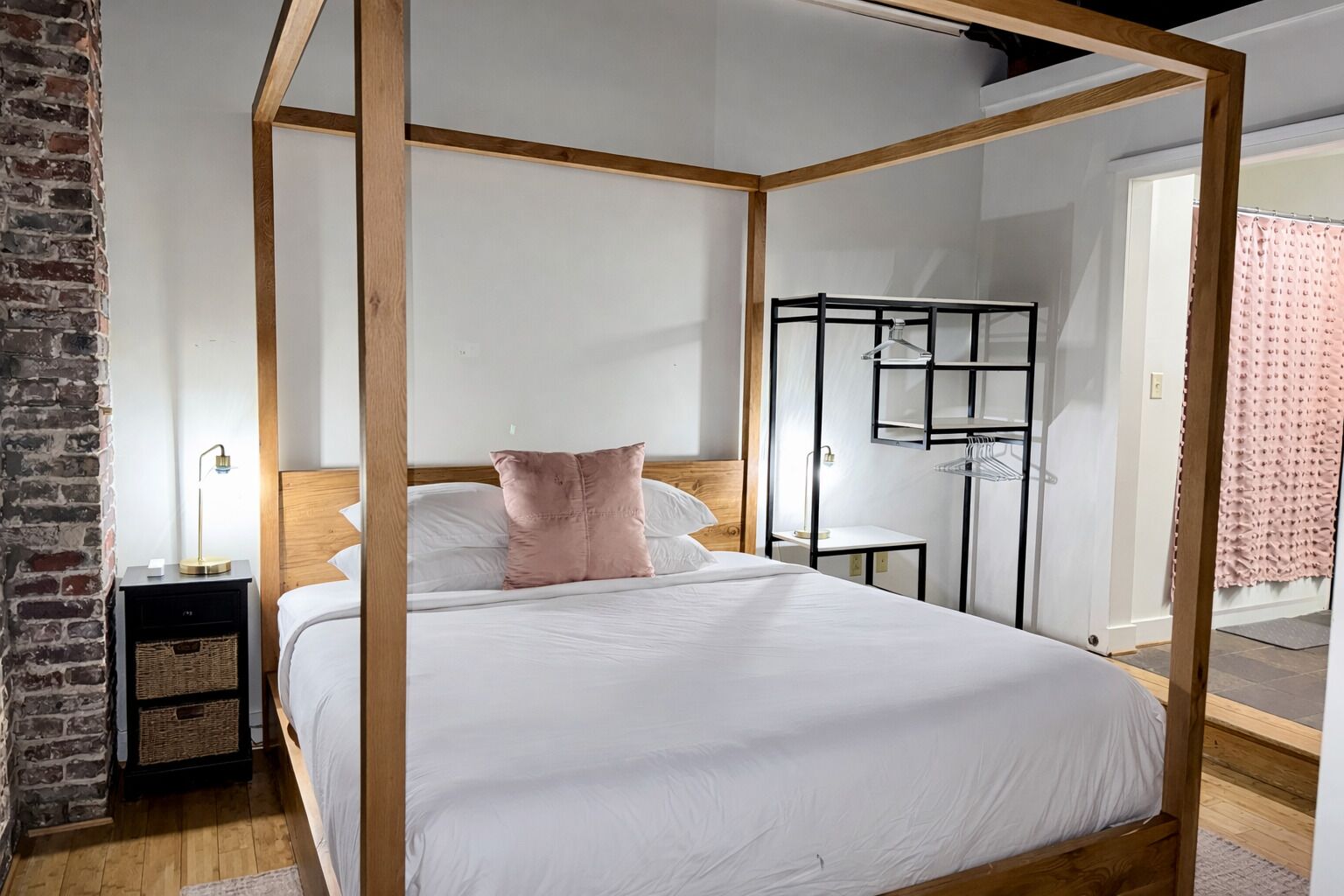 Una cama con dosel de madera, ropa de cama blanca y una única almohada rosa. Hay una mesita de noche con una lámpara y una cesta de mimbre. Se ve un armario metálico negro al fondo.