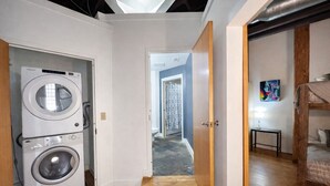 A laundry room with stacked washers and dryers, a wooden staircase leading to a hallway, and a bunk bed in the adjacent room. - Downtown Music City Loft living! ~Steps to the Ryman and Broadway (Nashville)