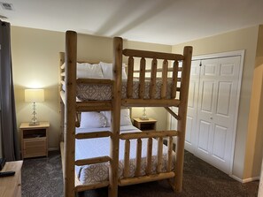 Una cama litera con estructura de madera, una mesita de noche con lámpara, y una puerta al fondo.