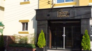 Un bâtiment avec une enseigne indiquant « AUBERGE ESPACE » à l’entrée, doté d’un design moderne et agrémenté de deux arbres à feuilles persistantes en pot devant l’entrée.