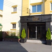 A building with a sign reading "HOSTEL ESPACE" at the entrance, featuring a modern design with two potted evergreen trees in front.