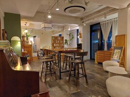 A cozy cafe with a wooden bar, high chairs, a bookshelf, and a large window with curtains.