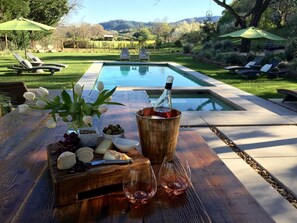 Escena junto a la piscina con una mesa de madera que presenta una tabla de quesos, uvas, aceitunas y una botella de vino.