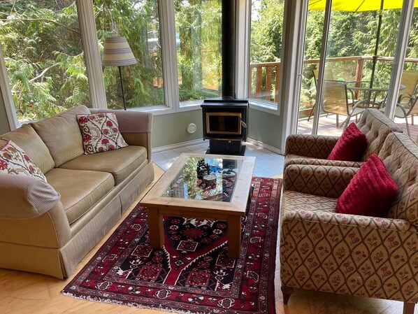 A cozy living room with a sofa, armchair, and a glass coffee table. A fireplace is visible in the corner, and the room has large windows offering a view of greenery outside.