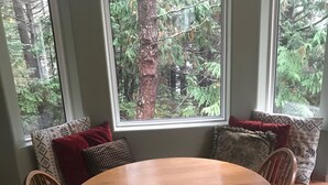Une table de salle à manger ronde en bois, accompagnée de quatre chaises, placée près d’une grande fenêtre donnant sur une forêt.