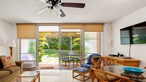 Un vaste espace de vie comprenant un canapé, une table basse et des meubles en osier. Une porte-fenêtre coulissante donne accès à un patio équipé d’une table et de chaises. On peut également apercevoir un ventilateur de plafond et un téléviseur à écran plat.