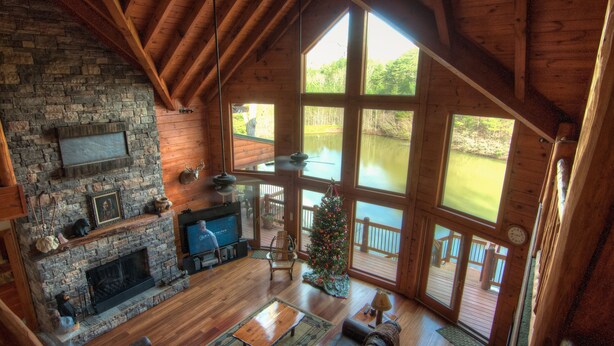 A spacious living room with a stone fireplace, wooden beams, a large window overlooking a lake, and a Christmas tree.