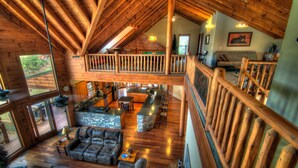 A spacious loft-style living area with a high ceiling, wooden beams, a stone fireplace, a leather sofa, and a dining area with a bar.