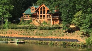 A large wooden cabin with a balcony, situated by a lake surrounded by dense trees.