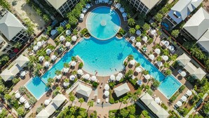 Une grande piscine centrale, entourée de chaises longues et de parasols, avec des palmiers et un sol à motifs à carreaux.