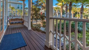 Une terrasse en bois avec un banc et un paillasson, entourée d’une balustrade blanche et de palmiers.